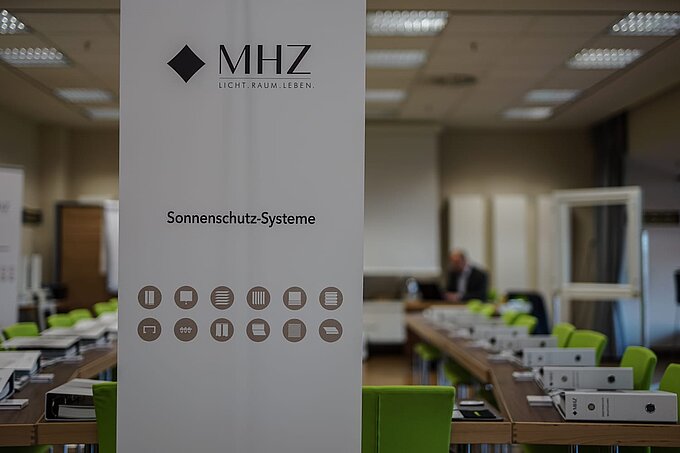MHZ-Roll-up „Sonnenschutz-Systeme“ in einem Seminarraum mit grünen Stühlen und Ordnern.