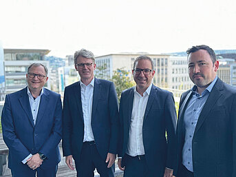 Vier Männer im Anzug posieren auf einer Dachterrasse vor Bürogebäuden