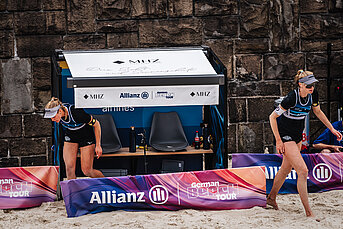 Zwei Beachvolleyball-Spielerinnen stehen vor einer schattenspendenden MHZ-Markise mit Sitzbank beim Turnier der German Beach Tour.