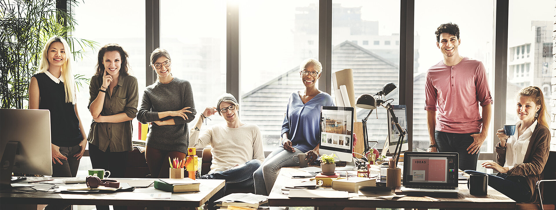 Gruppe von Mitarbeitern in einem modernen Büro mit großen Fenstern und viel Tageslicht, umgeben von Arbeitsplätzen.