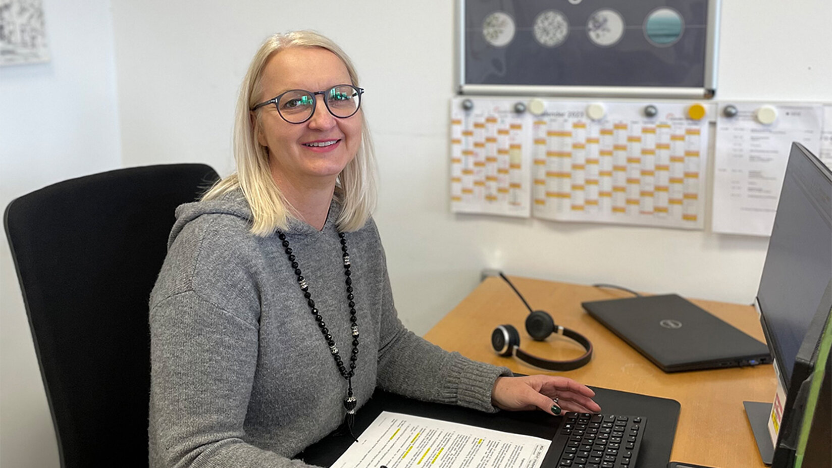 Frau mit Brille sitzt an einem Schreibtisch und arbeitet am Laptop, im Hintergrund ein Kalender an der Wand.