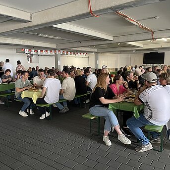 MHZ-Mitarbeitende sitzen beim Sommergrillen 2024 in einer Halle an langen Tischen, essen, trinken und unterhalten sich in geselliger Atmosphäre.