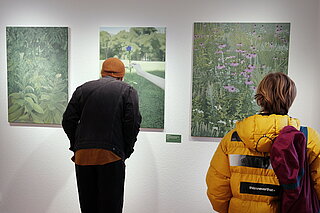 Zwei Personen betrachten in einer Galerie drei an der Wand hängende Kunstwerke mit grünen, naturähnlichen Motiven. Eine Person trägt eine gelbe Jacke, die andere eine dunkle Kleidung und Mütze.
