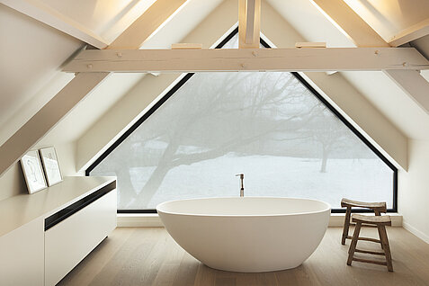 Badezimmer mit freistehender Badewanne vor einem großen Dreiecksfenster mit Sichtschutztextil.