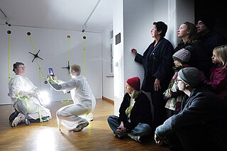 In einem Ausstellungsraum führen zwei Personen in weißen Anzügen eine Performance mit hellen Lichtquellen auf, während mehrere Zuschauer an der Wand sitzen und die Szene beobachten.