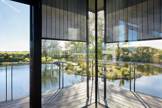 Innenansicht mit bodentiefen Glasflächen und teilweise abgesenkten außenliegenden Screen Sonnenschutzanlagen mit Blick auf eine Terrasse und einen naturgestalteten Teich.