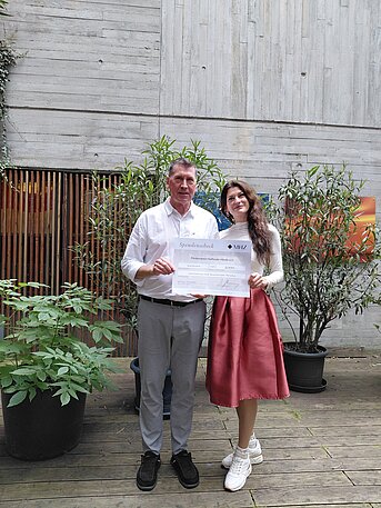 Zwei Personen stehen in einem begrünten Innenhof und halten gemeinsam einen Spendenscheck als Zeichen sozialen Engagements eines Unternehmens.