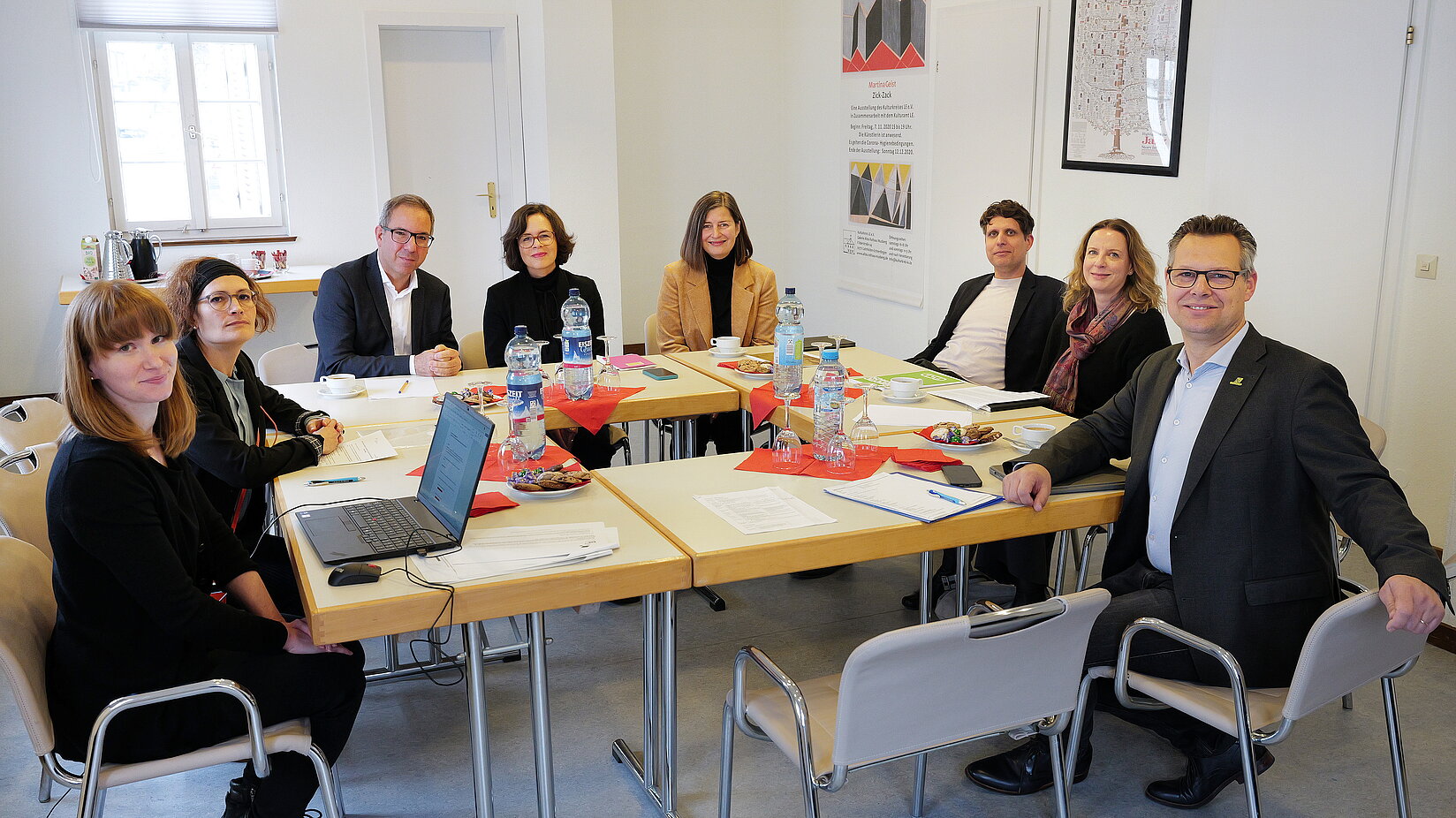 Alternativtext: Mehrere Personen sitzen in einem Besprechungsraum an einem rechteckigen Tisch mit Unterlagen, Laptops und Getränken. Alle blicken in Richtung Kamera. An der Wand hängen Bilder und Grafiken.