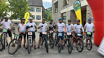 ehn Radfahrer in weißem MHZ-Trikot mit Startnummern stehen nebeneinander mit ihren Mountainbikes unter einem roten Torbogen.