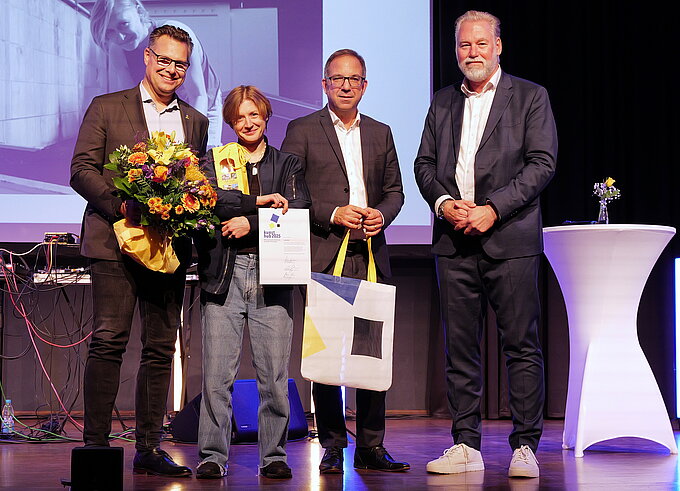 Vier Personen stehen auf einer Bühne. Eine Person hält einen Blumenstrauß und eine Urkunde, daneben stehen zwei Männer mit Geschenktüten und eine weitere Person neben einem weißen Stehtisch. Im Hintergrund ist eine Leinwand mit Projektion zu sehen.