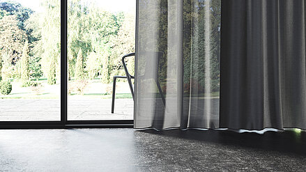 Nahaufnahme eines weißen MHZ-Stores und eines grauen Vorhangs vor einem großen Fenster mit Blick ins Grüne.