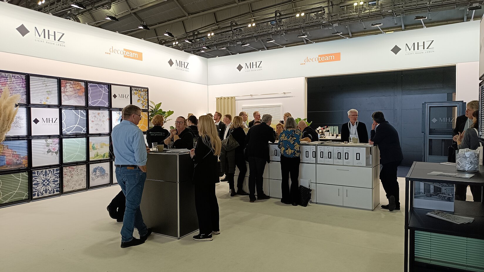 Besucher im Gespräch mit MHZ-Mitarbeitenden am Messestand mit Plissee-Präsentation und Theke auf einer Fachmesse.