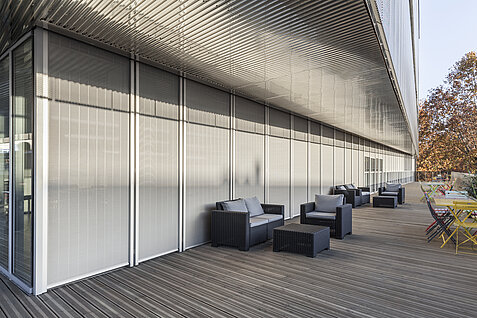 Terrasse mit Sitzmöbeln vor einer Glasfassade mit außenliegenden vertikalen Senro Metallbehängen als Sonnenschutz.
