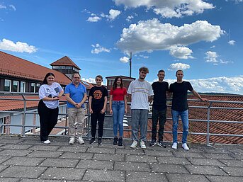 Sieben Auszubildende stehen bei Sonnenschein auf einer Dachterrasse mit Blick auf Ziegeldächer und blauen Himmel.