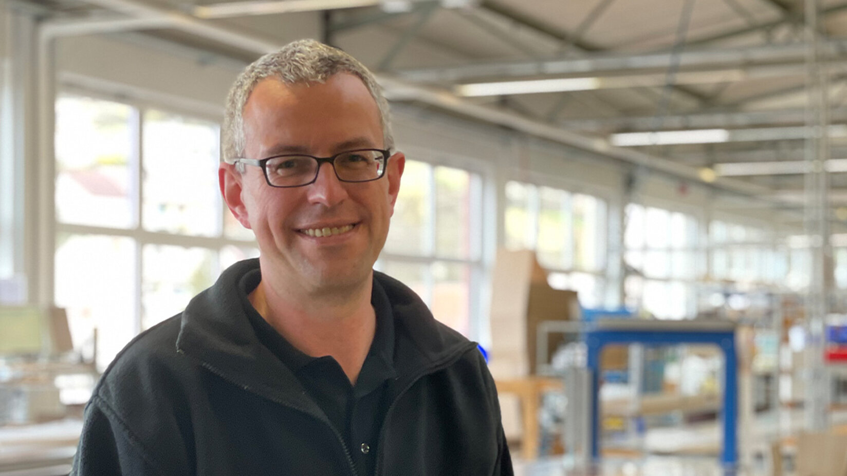 Mann mit Brille steht lächelnd in einer hellen Produktionshalle mit großen Fenstern im Hintergrund.