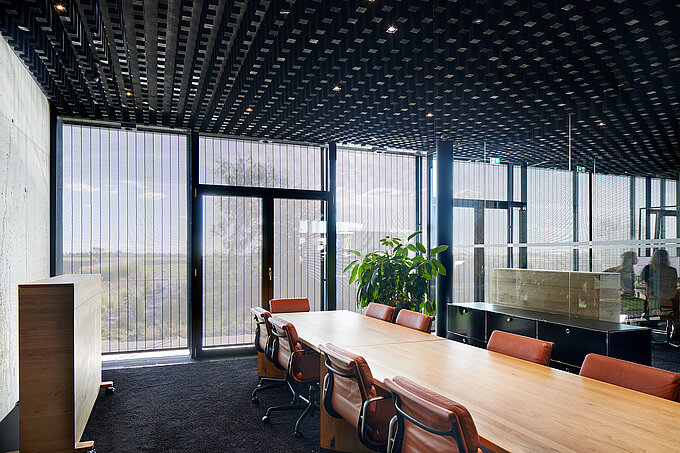 Moderner Konferenzraum mit großer Fensterfront und außenliegendem Metallbehang s_enro, der Tageslicht filtert und effektiven Sicht- und Sonnenschutz bietet.