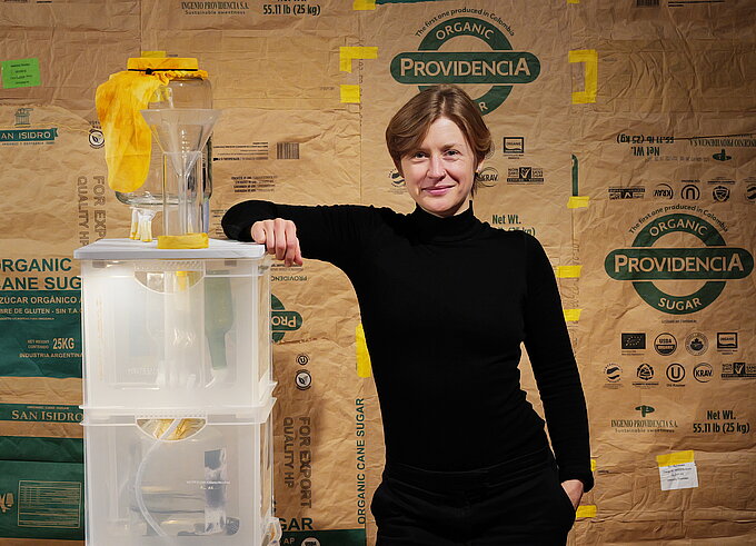 Eine Frau steht neben einer Skulptur aus transparenten, übereinandergestapelten Würfeln mit einem gelben Element an der Spitze. Im Hintergrund sind Verpackungsmaterialien der Marke „Rockpanel“ zu sehen.