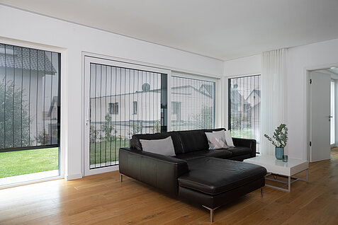 Helles Wohnzimmer mit großem Fenster, schwarzem Ledersofa und MHZ Metallbehang sonro als Sicht- und Sonnenschutz.