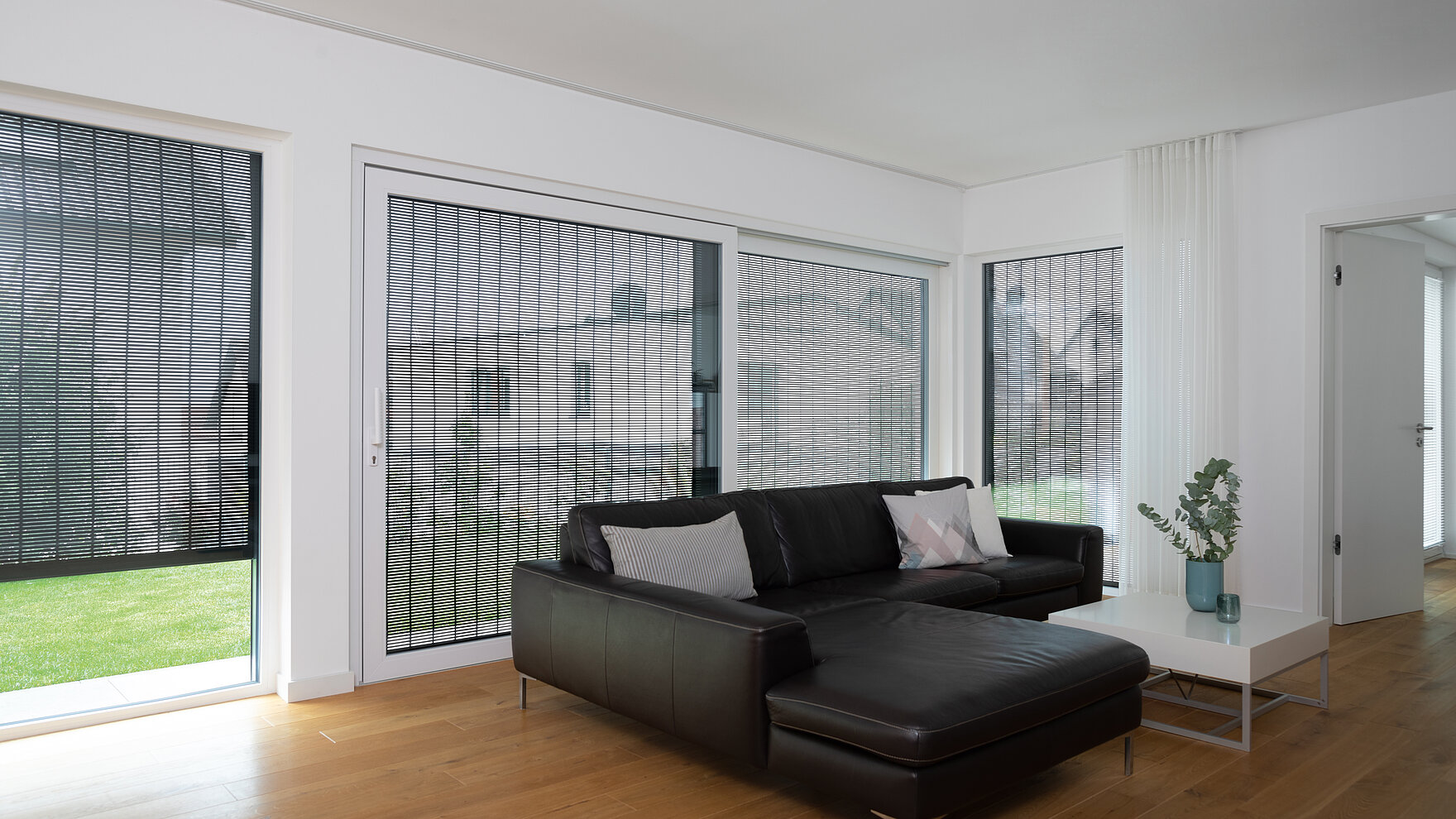 Bright living room with large windows, black leather sofa, and MHZ sonro metal blinds as sun and privacy protection.