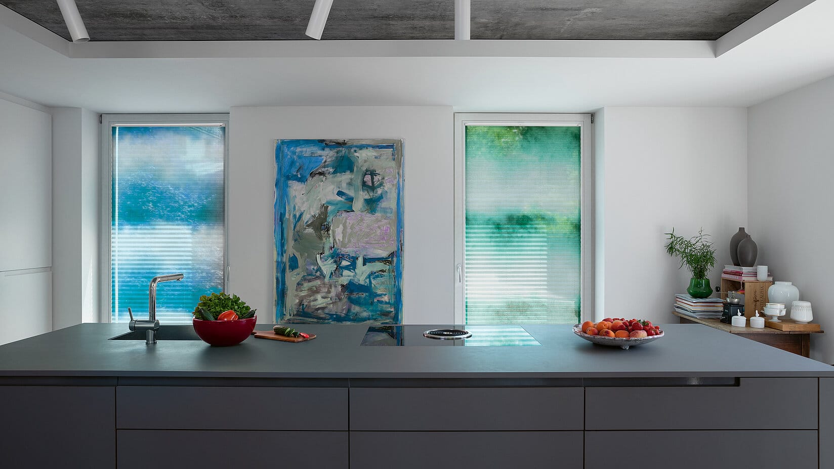 Küche mit Kücheninsel und zwei Fenstern die mit Plissees in Aquarelloptik in jeweils grün und blau vor Sonne schützen.