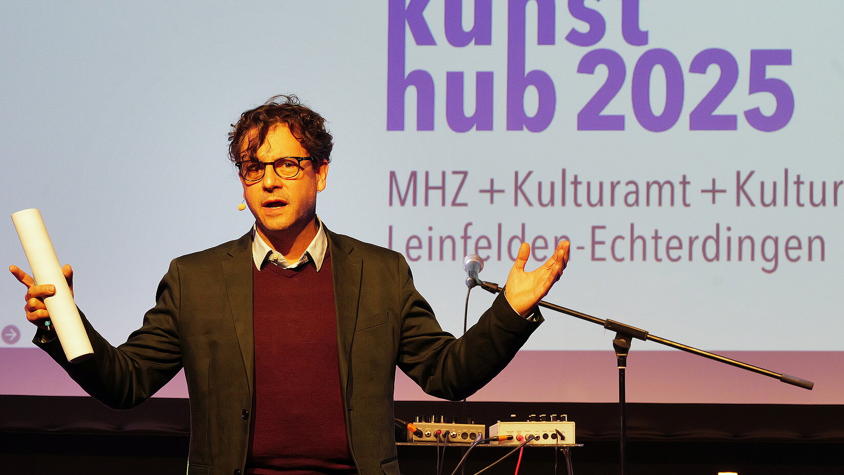Eine Person steht auf einer Bühne vor einer Leinwand mit der Aufschrift „kunsth ub 2025 – Kulturamt Leinfelden-Echterdingen“. Die Person hält ein Blatt Papier in der Hand und spricht zum Publikum. Im Hintergrund sind Musikinstrumente zu sehen.