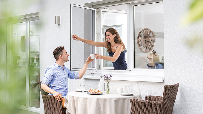 Woman hands man a cup through an open pivoting window with insect screen.
