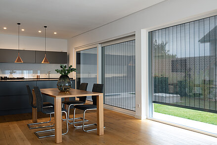 Moderne Wohnküche mit großen Fenstern und außenliegendem MHZ Metallbehang Sonro, Holztisch im Vordergrund.