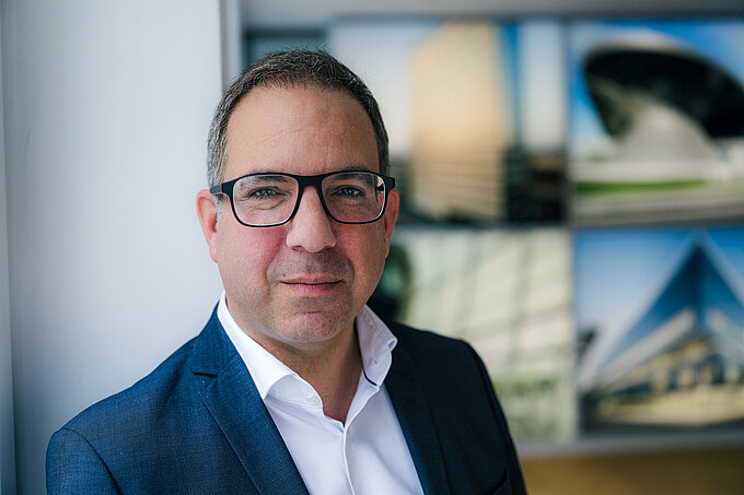 Man in a suit sitting with a laptop at a table in a bright meeting room with large windows.