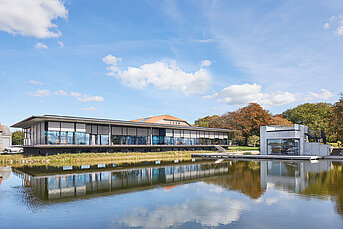 Moderne Glasfassade mit außenliegenden Screen Sonnenschutzanlagen an einem großen Wasserbecken mit klarer Spiegelung und herbstlicher Landschaft im Hintergrund.