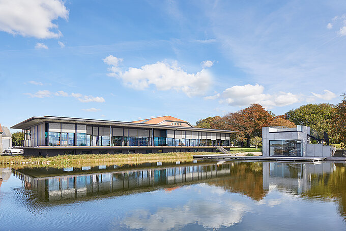 Moderne Glasfassade mit außenliegenden Screen Sonnenschutzanlagen an einem großen Wasserbecken mit klarer Spiegelung und herbstlicher Landschaft im Hintergrund.