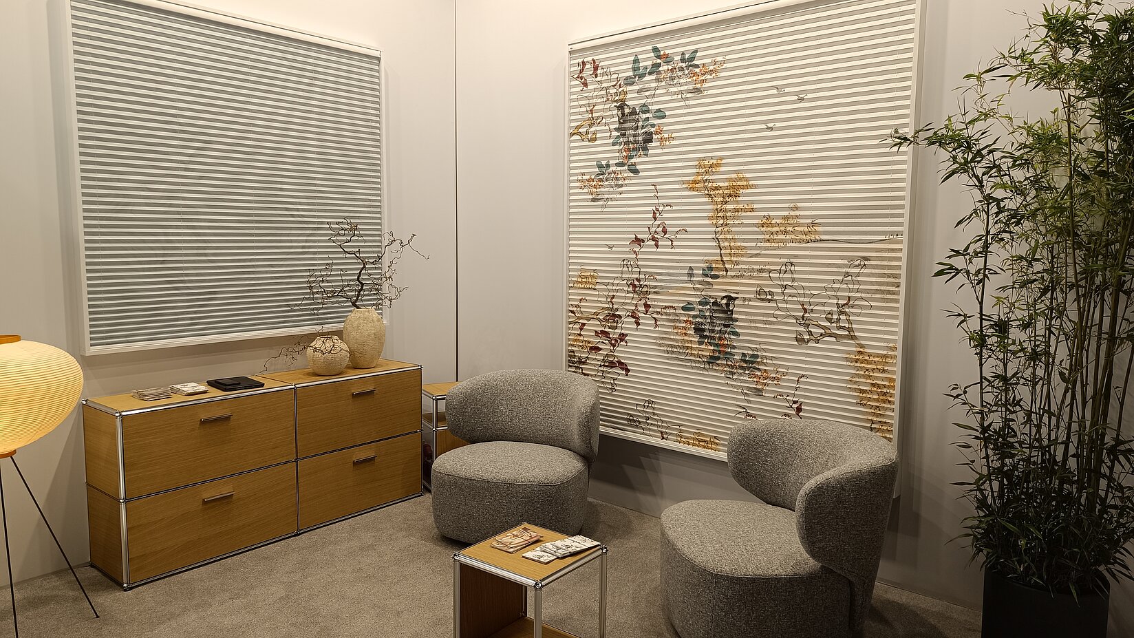 Wohnraum mit zwei Fenstern und Plissees, eines davon mit dekorativem Pflanzenmotiv als Sicht- und Sonnenschutz.