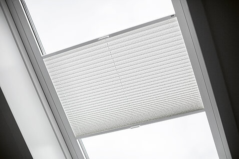 Weißes Plissee in einem Dachflächenfenster mit lichtdurchlässigem Stoff zur Lichtregulierung.