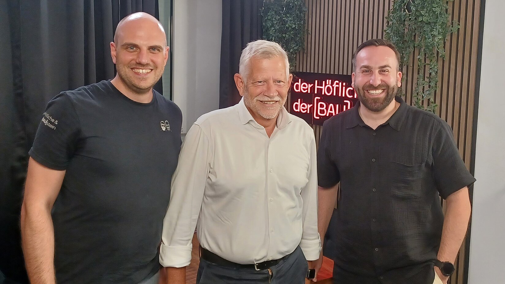 Drei Männer stehen nebeneinander in einem Podcast-Studio und lächeln in die Kamera.