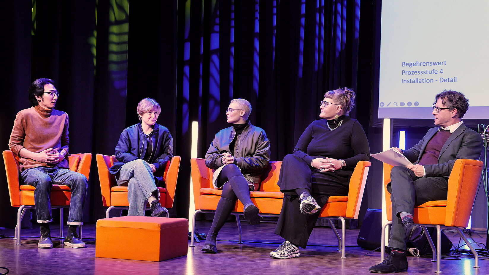 Fünf Personen sitzen auf einer Bühne in orangefarbenen Sesseln und führen eine Podiumsdiskussion. Eine Person moderiert das Gespräch. Im Hintergrund sind dunkle Vorhänge und eine Projektionsfläche zu sehen.