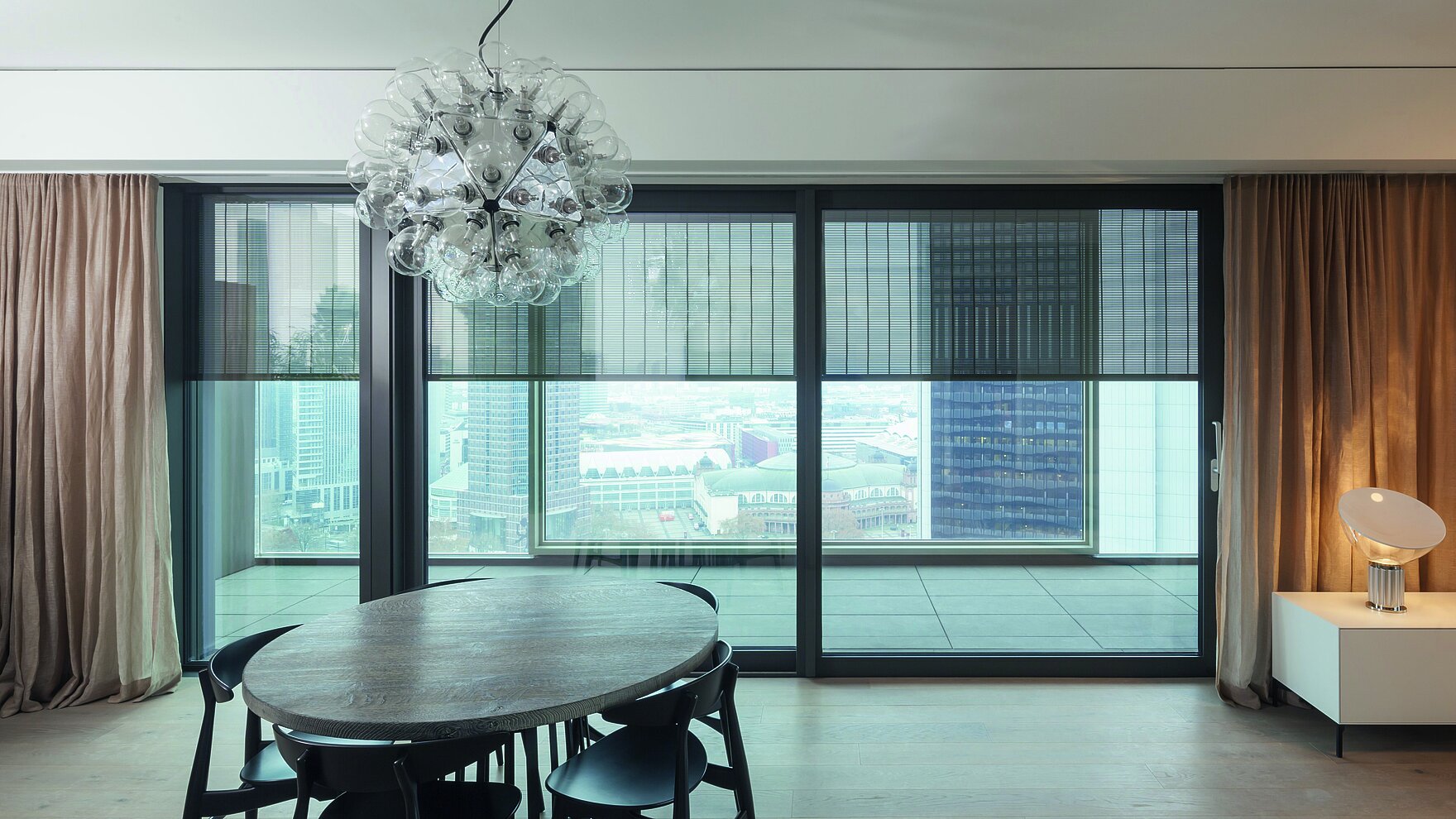 View from a modern living area with dining table toward large windows fitted with external SENRO metal blinds, with city view outside.