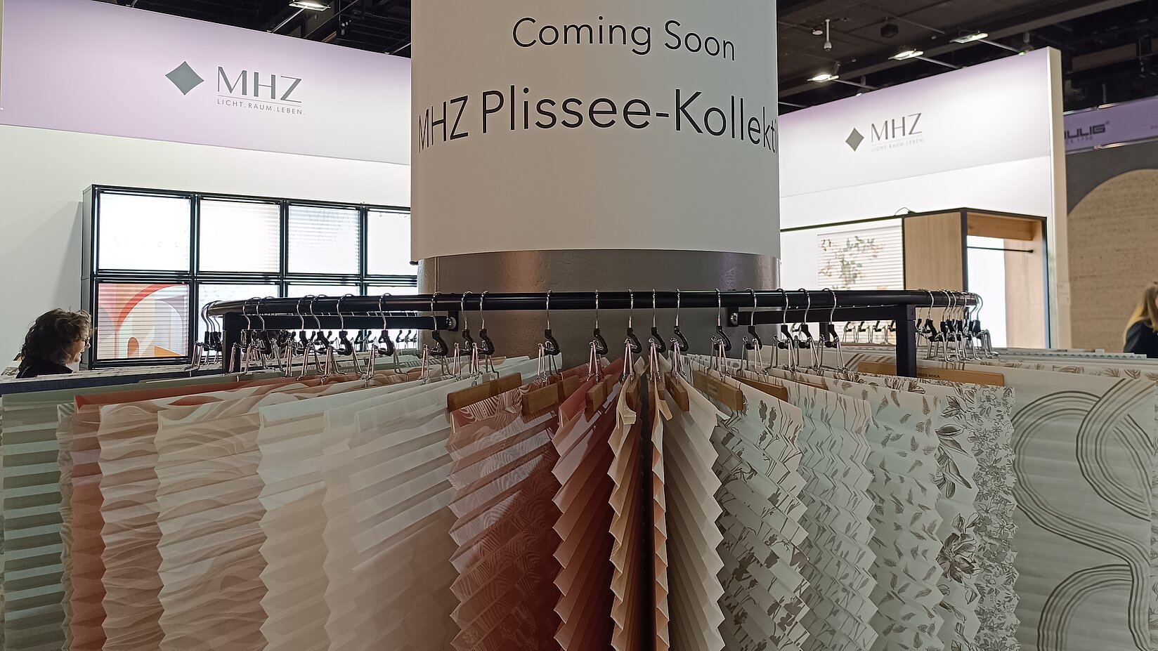 Messestand von MHZ mit aufgehängten Plissee-Stoffmustern und dem Hinweis „Coming Soon – MHZ Plissee-Kollektion“.
