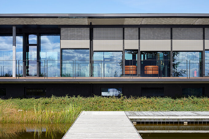 Moderne Gebäudefassade mit großen Glasflächen und außenliegenden Screen-Sonnenschutzanlagen vor einer Terrasse mit Glasgeländer und Holzsteg.
