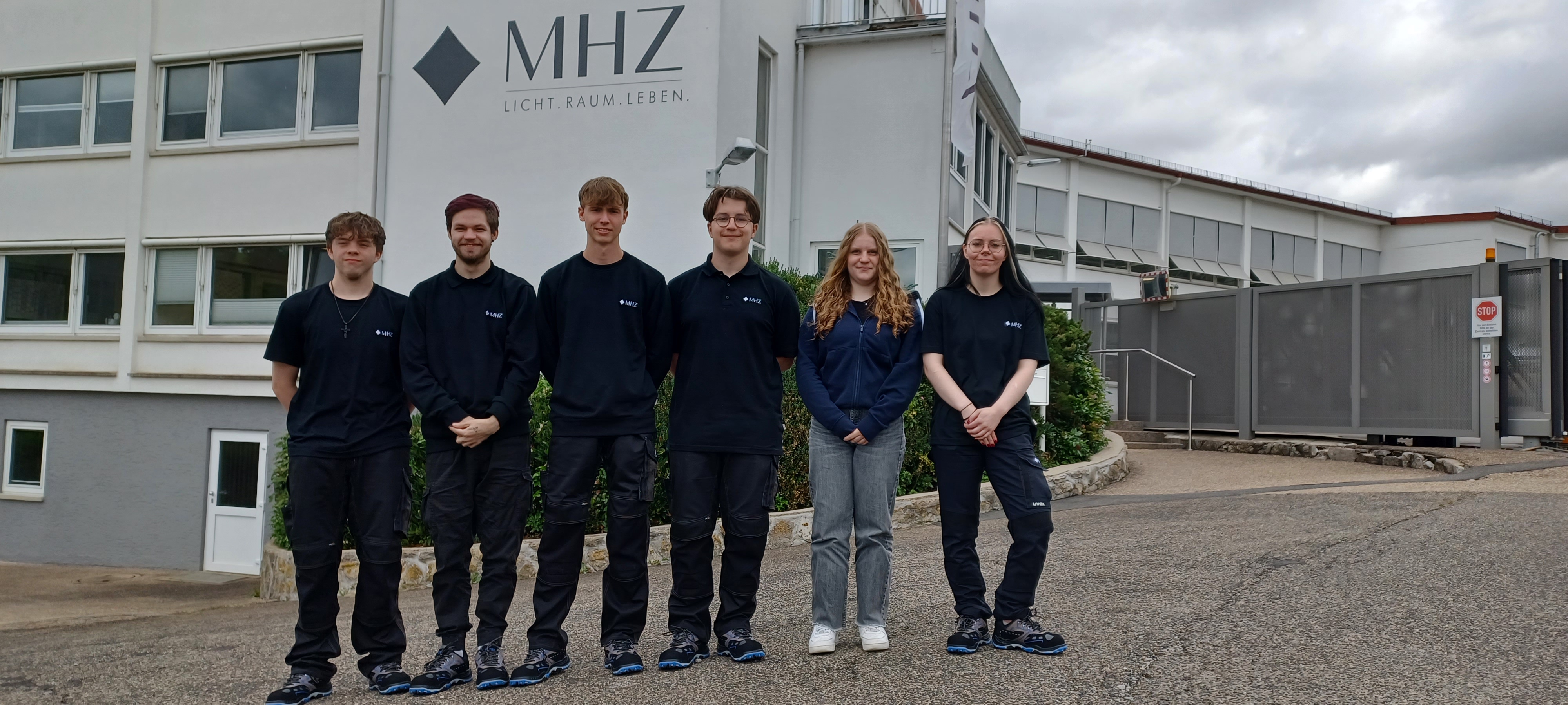Sechs junge Mitarbeitende der Firma MHZ stehen vor dem Firmengebäude in Arbeitskleidung mit Logo. 