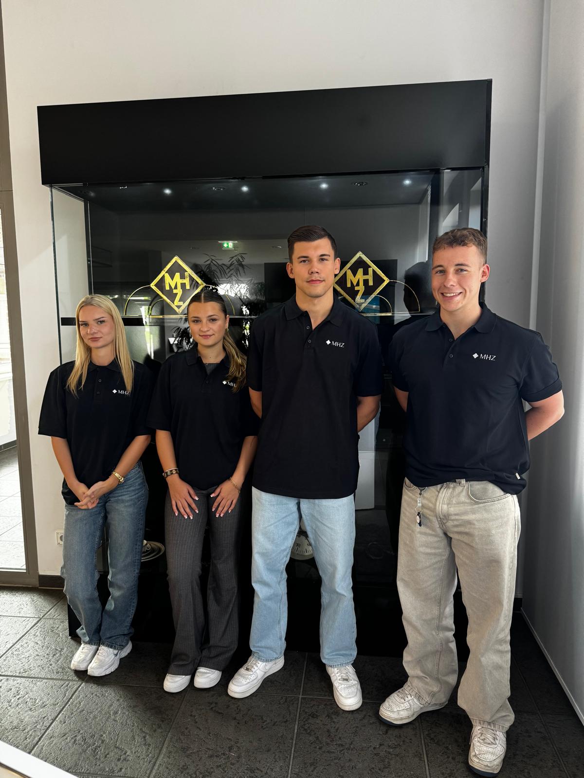 Vier junge Mitarbeitende von MHZ stehen gemeinsam vor einer Glasvitrine und tragen schwarze Poloshirts mit Firmenlogo.